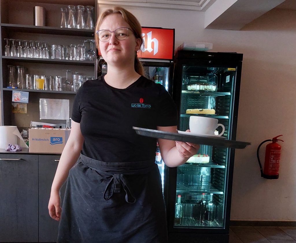 Eine junge Frau hält ein Tablett in der Hand. Ihr Blick wirkt selbstbewusst. Sie hat eine Schürze umgebunden und trägt ein schwarzes Oberteil. Offensichtlich arbeitet sie in einem gastronomischen Betrieb.