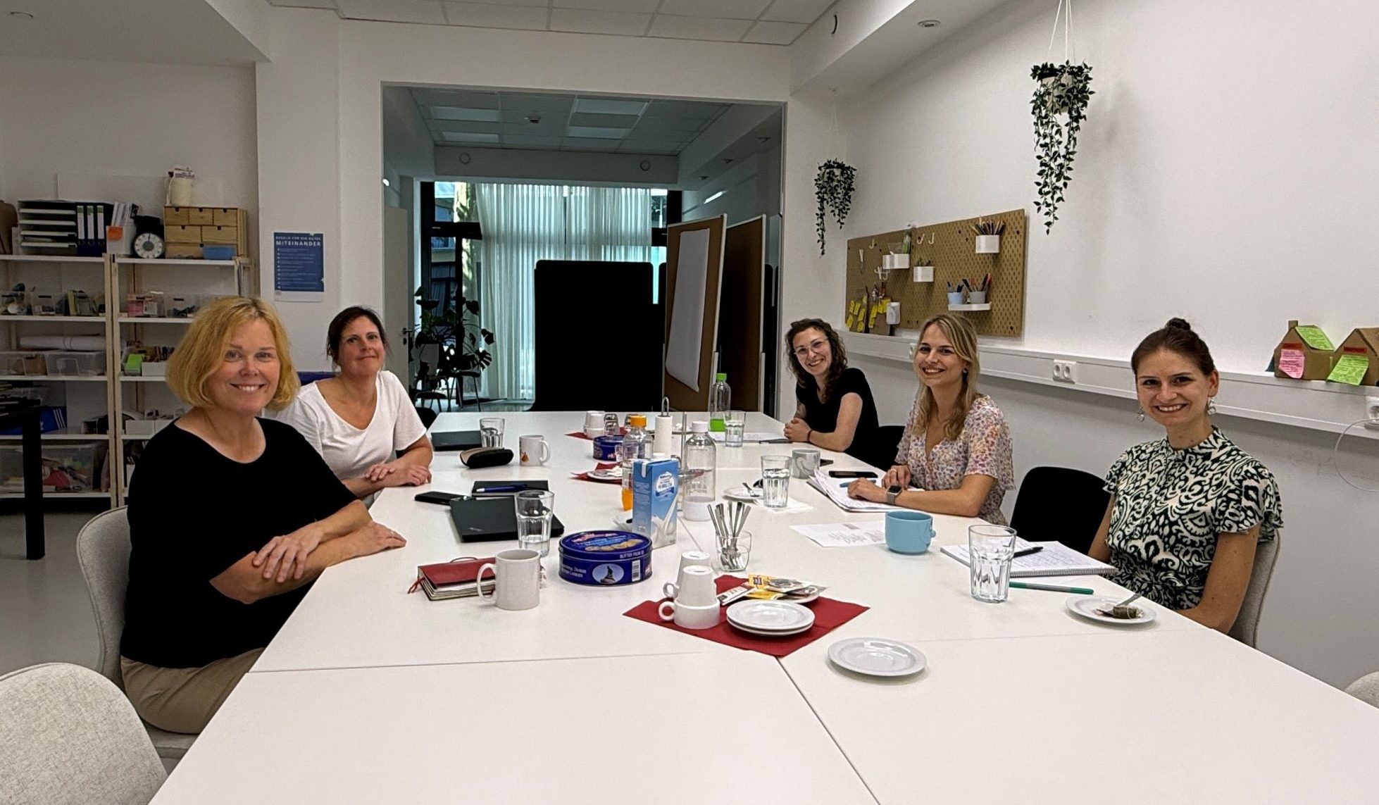 Eine Gruppe von fünf Frauen sitzt um einen langen Arbeitstisch. Sie blicken zur Kamera und wirken gut gelaunt. Im Hintergrund sind unter anderem Pinnwände und Flipcharts zu erkennen. Wir befinden uns offenbar in einem Tagungsraum.