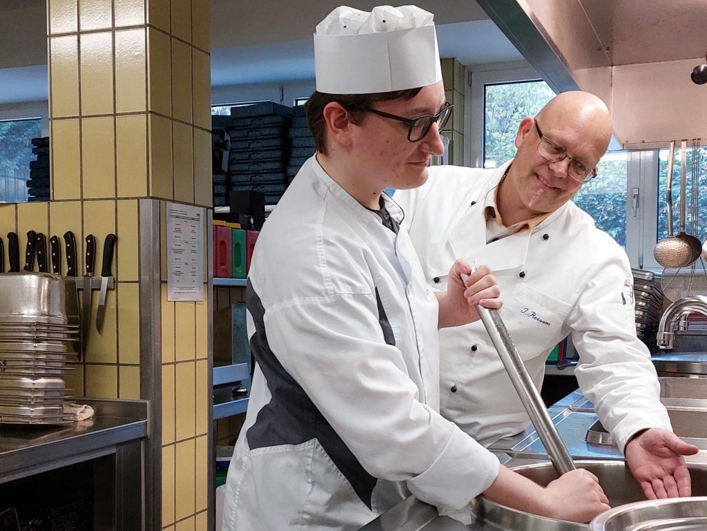 Zwei Männer stehen in einer Großküche. Einer der beiden hält ein großes Küchengerät in der Hand und rührt in einem Topf, der zweite deutet mit seiner linken Hand auf den Topf.