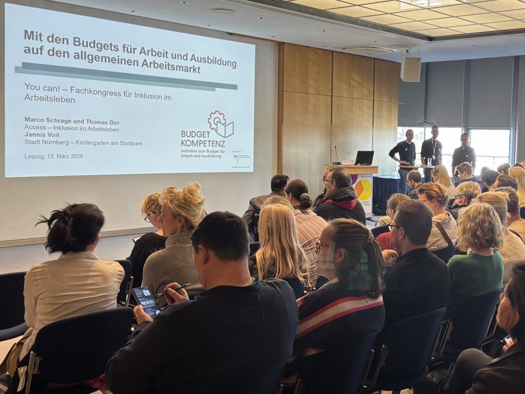 Eine Vortragsszene: Drei Personen stehen vorne, davor sind viele Zuhörende zu sehen.