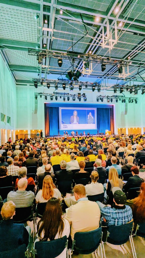 Ein voll besetzter Veranstaltungssaal. Der Saal ist in grünliches Licht getaucht. Die Bühne ist weit entfernt, über der Bühne ist ein großer Monitor.