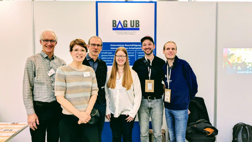 Eine Gruppe von Personen steht vor dem Stand der BAG UB und lacht.