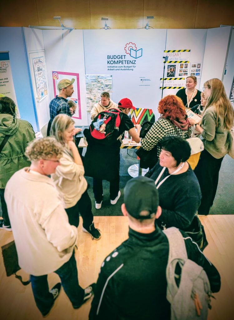 Eine Gruppe von Menschen steht um den Infostand des Projektes Budgetkompetenz.