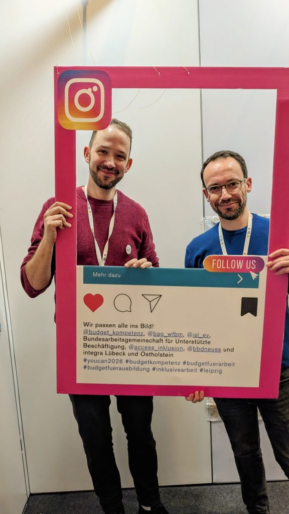 Zwei Personen schauen durch einen pinken Rahmen. Auf dem Rahmen steht: Wir passen alle ins Bild.