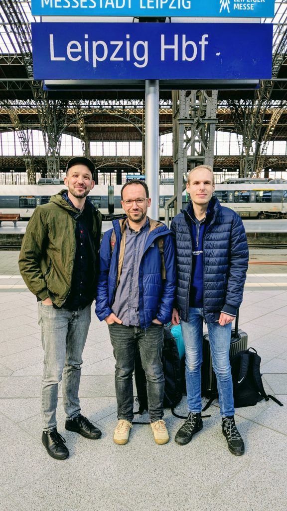 Drei Männer stehen am Hauptbahnhof von Leipzig am Gleis. Sie lächeln in die Kamera.