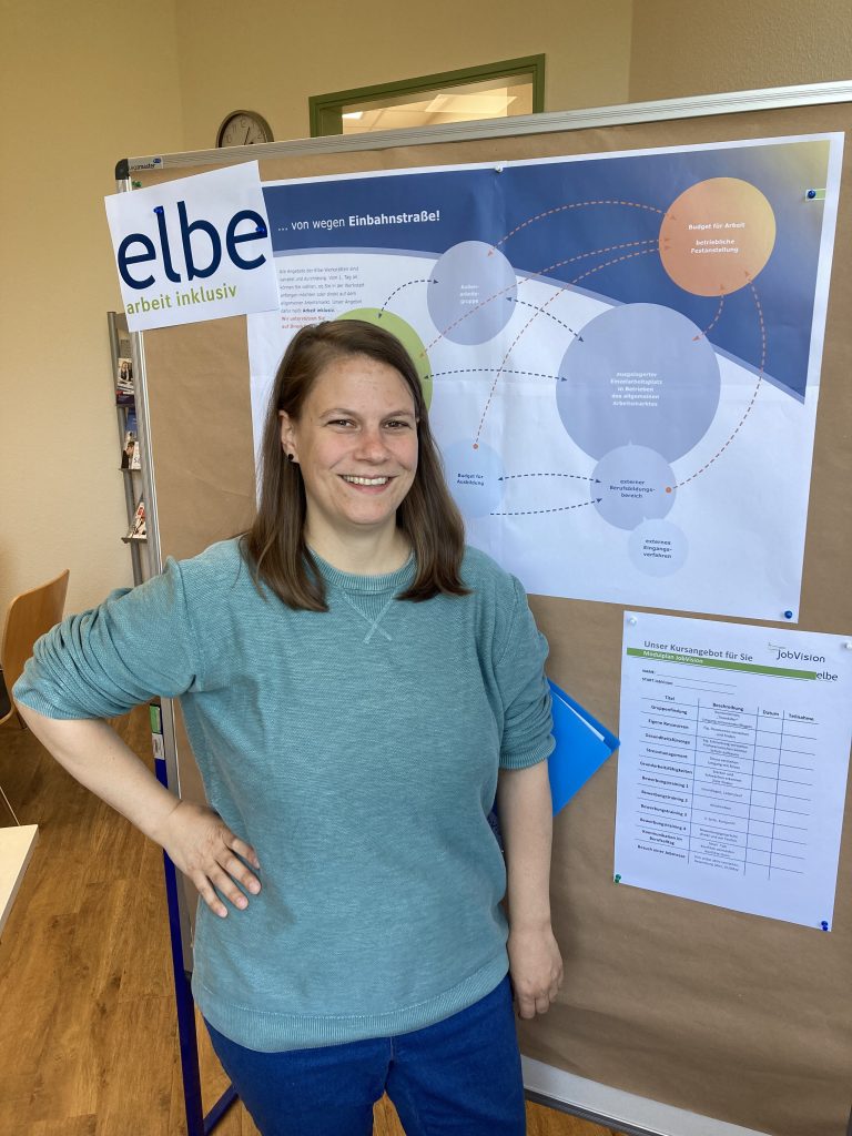 Eine Frau in einem blau-grauen Pullover steht vor einer Pinnwand. Auf der Pinnwand ist zu lesen: elbe arbeit inklusiv